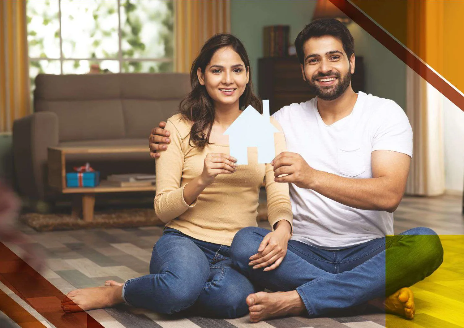 A young couple sitting on the floor of their living room, holding up a paper cut-out of a house